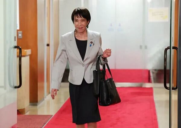 A woman in a light jacket with two black bags over her left arm smiles as she walks on red carpet through a doorway.