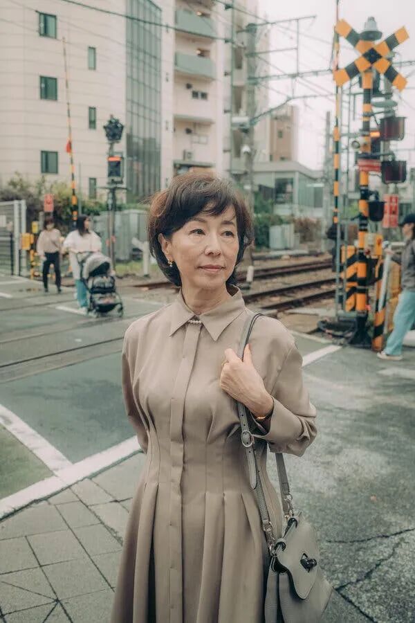 A woman in a long, light-brown dress stands at a railway crossing on the street.