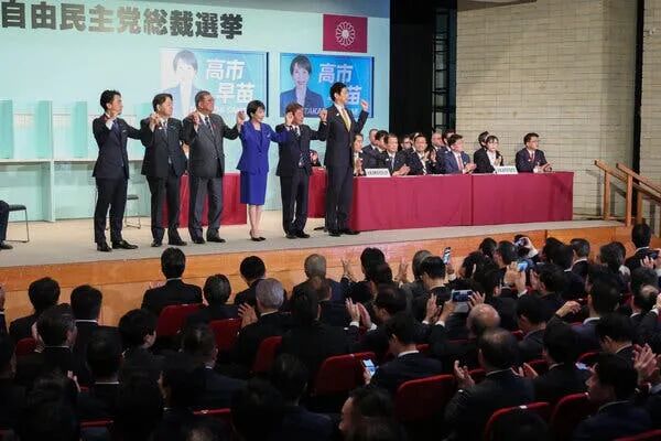 A woman in a  blue suit stands on a stage holding hands aloft with men in suits alongside her.