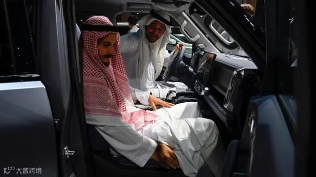 在一场车展上,两名身穿长袍、头戴头巾的男子,分别坐在一辆中国制造的汽车的驾驶座位和前排乘客座位。