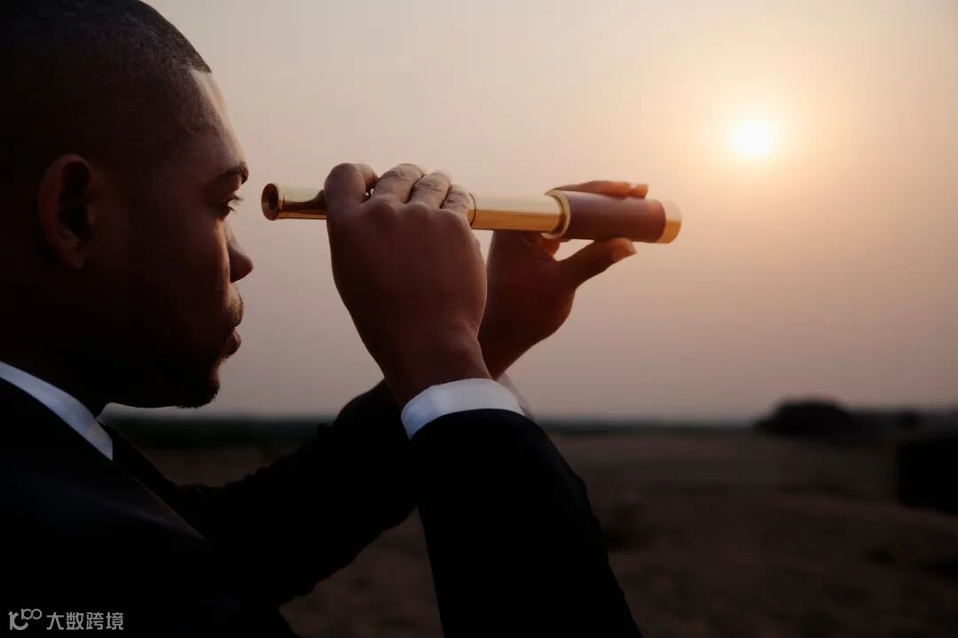 A man in a business suit looking through a telescope toward the horizon.