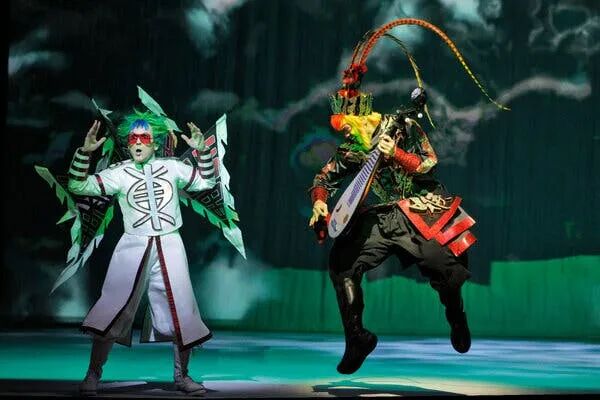 A man with green hair in a vibrant cartoony costume sings on the opera stage; beside him is a dancer slightly off the ground. He has monkey plumage and holds a string instrument.