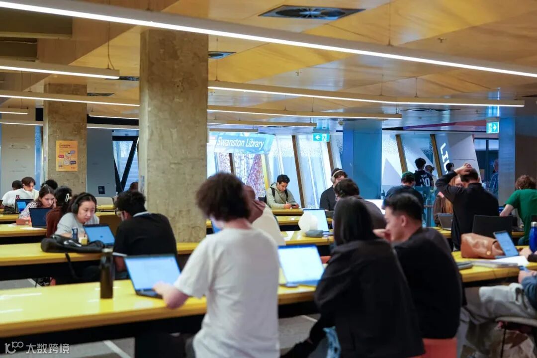 University students sitting in front of laptops on long wooden desks in a hall.