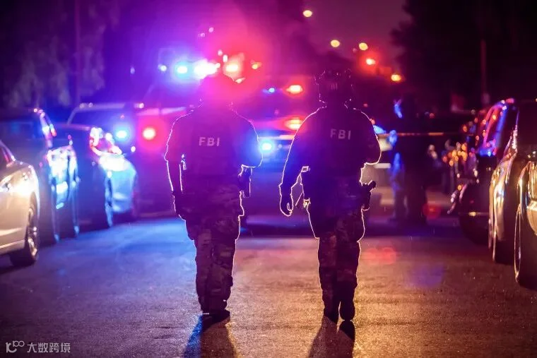 Two FBI field agents wearing helmets and camouflage fatigues walk in front of a vehicle with red and blue flashing lights.
