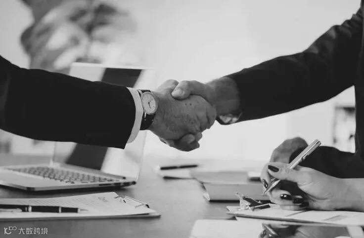 This may contain: two men shaking hands over a desk with laptops and papers in front of them