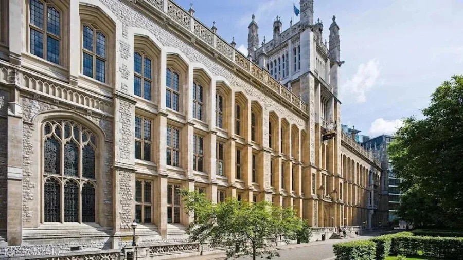 A university library which resembles a large chapel with a tower in the middle. There is a hedge and a tree on the right and a sapling in the foreground.
