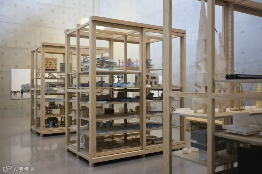 Light wood display shelves filled with architectural models, drawings, and material samples in a spacious gallery with concrete walls.